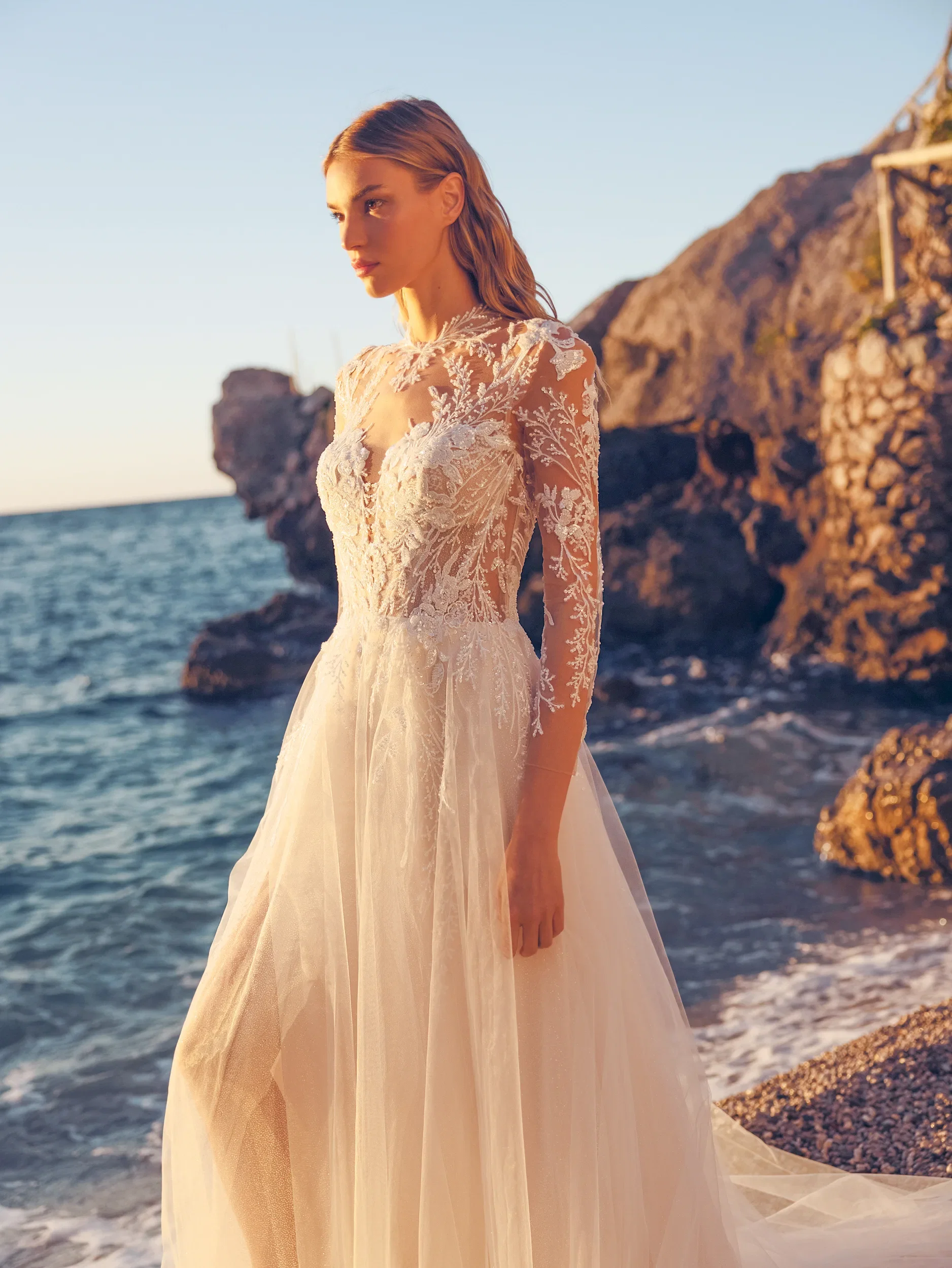 Bruid in trouwjurk met prachtig kant en tule. Op de achtergrond is de zee met rotsen te zien.