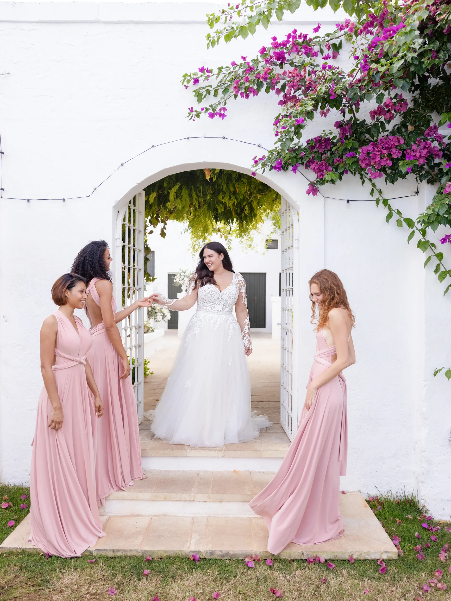 Vrolijke bruid in kanten bruidsjurk met tule, omringd door haar bruidsmeisjes in roze jurken.