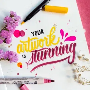 Kleurrijk kunstwerk met handlettering in geel en roze, met stiften en bloemen op flatlay.