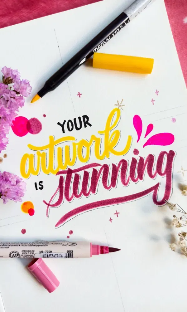 Kleurrijk kunstwerk met handlettering in geel en roze, met stiften en bloemen op flatlay.