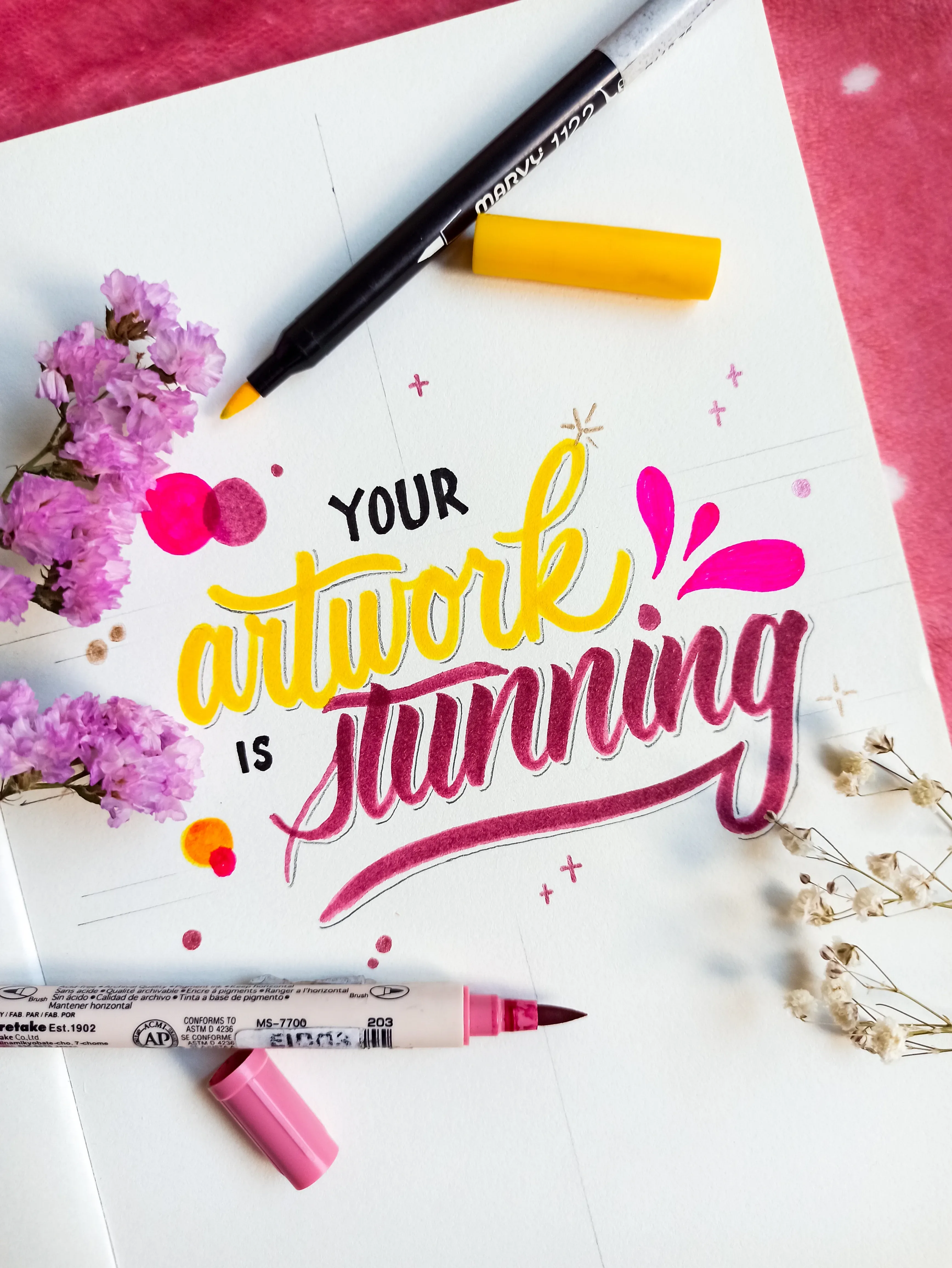 Kleurrijk kunstwerk met handlettering in geel en roze, met stiften en bloemen op flatlay.