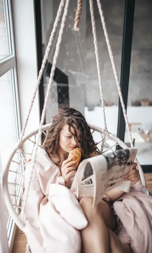 Vrouw ontspant op een zitstoel met een croissant en een krant.