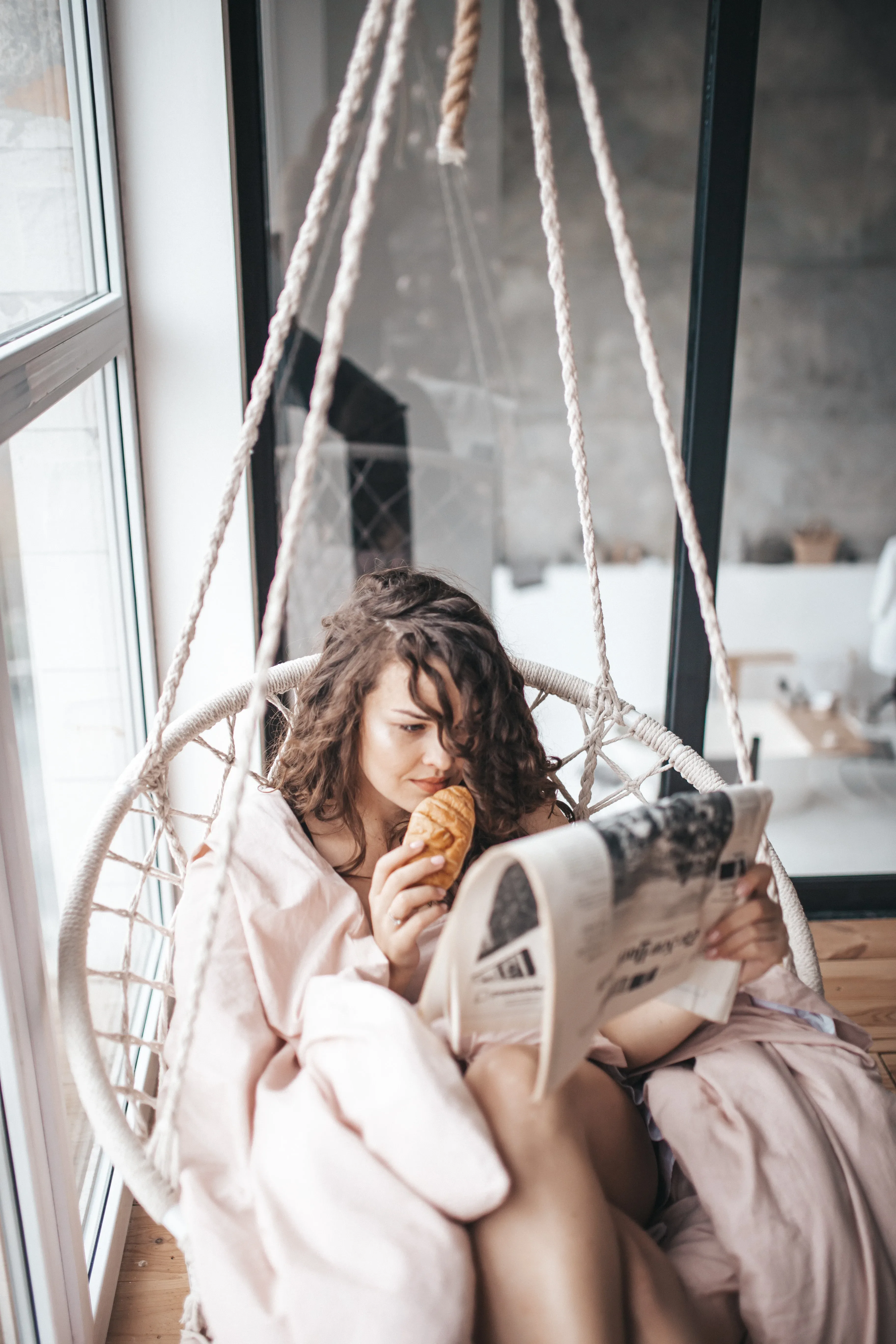 Vrouw ontspant op een zitstoel met een croissant en een krant.