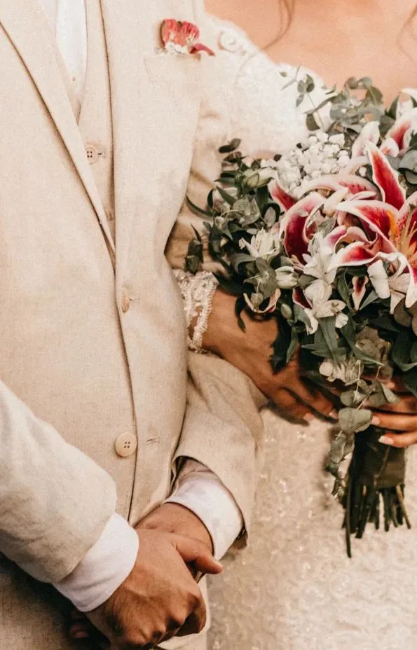 Bruidspaar staat gearmd naast elkaar. Bruid houdt een bruidsboeket vast. Bruidegom draagt zandkleurig driedelig trouwpak.