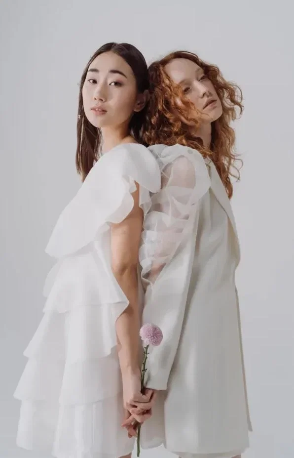 Twee vrouwen staan met de ruggen tegenover elkaar. Beide handen houden een roze bloem vast