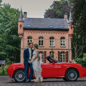 Bruidspaar voor rode cabrio trouwvervoer, met op de achtergrond een landhuis trouwlocatie.