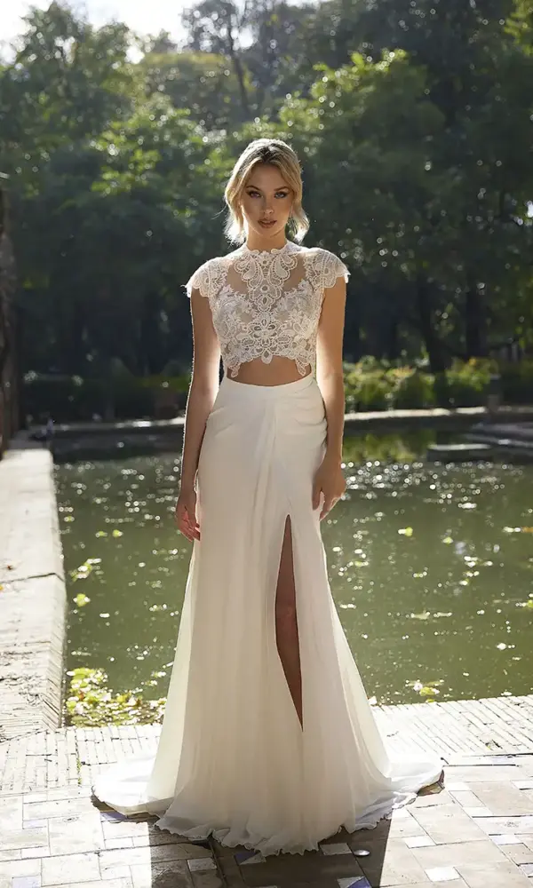 Een sierlijke vrouw in een unieke tweedelige trouwjurk staat langs een parkvijver. De top heeft een hooggesloten halslijn met delicate kanten details en korte mouwen, terwijl de lange rok een gewaagde split heeft. Het zonlicht schijnt door de bomen en reflecteert op het water, wat een heldere en natuurlijke ambiance creëert.