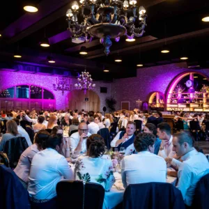 Grote zaal met sfeerverlichting, huwelijksdiner. Meerdere ronde tafels vol mensen.