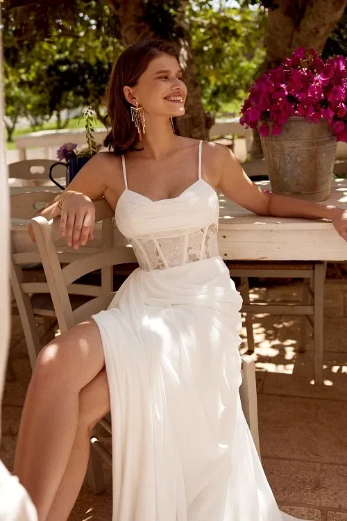 Vrouw in een moderne trouwjurk met spaghettibandjes, een transparant kanten korset en hoge split, ontspannen zittend aan een tafel in een zonovergoten tuin.