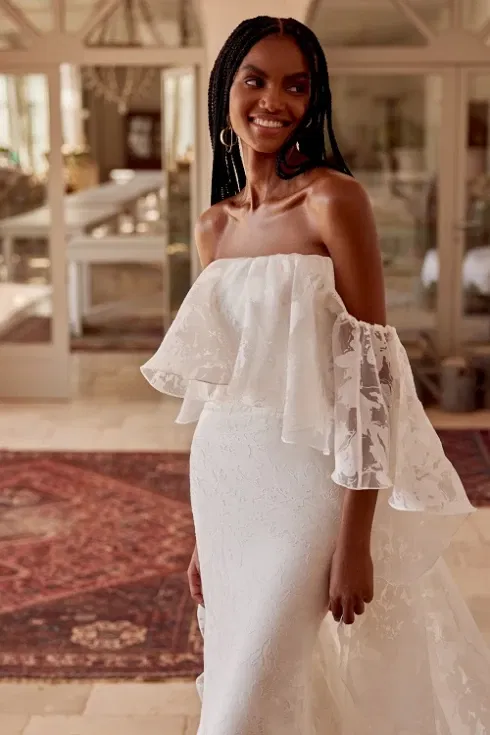 Lachende vrouw in een strapless trouwjurk met een romantisch bloemenreliëf en wijd uitlopende afneembare mouwen, poserend in een sfeervolle binnenruimte met oosterse tapijt.