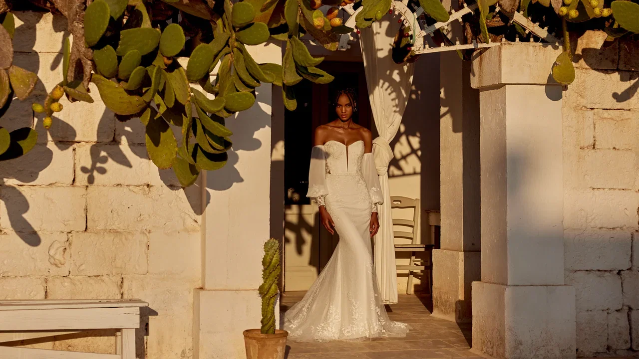 Vrouw in een elegante witte trouwjurk met off-shoulder pofmouwen en een lange sleep, staand in de deuropening van een mediterrane villa met zonlicht en cactusbladeren die schaduwen werpen op de muur.