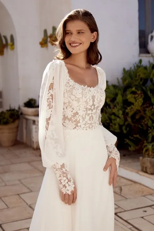 Vrouw in een elegante bohemian Lillian West trouwjurk met transparant kanten lijfje, pofmouwen en bloemenborduursels, staand op een zonovergoten terras met mediterrane beplanting.