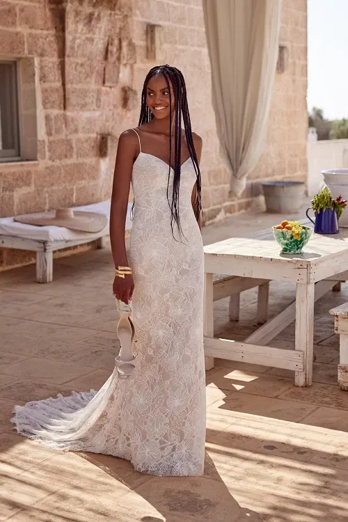 Vrouw in een elegante kanten trouwjurk van Lillian West met fijne spaghettibandjes en een sleep, poserend met haar rug naar de camera op een zonovergoten terras met rustieke stenen muren.