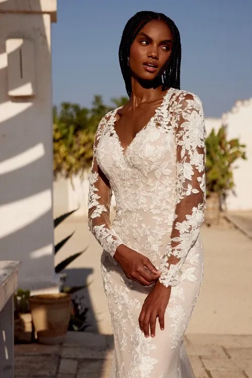 Bruid in een elegante trouwjurk met V-hals, transparante lange mouwen en witte bloemendetails, poserend in het zonlicht op een buitenlocatie.