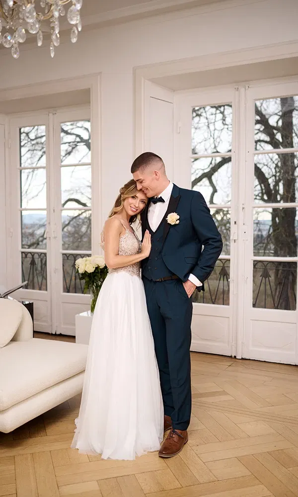 Bruid in een witte trouwjurk en bruidegom in donkerblauw pak, knus samen staand in een elegante kamer met grote ramen.