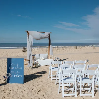 Ceremonieopstelling op een zonnig strand met witte stoelen, prieel en uitzicht op de zee.