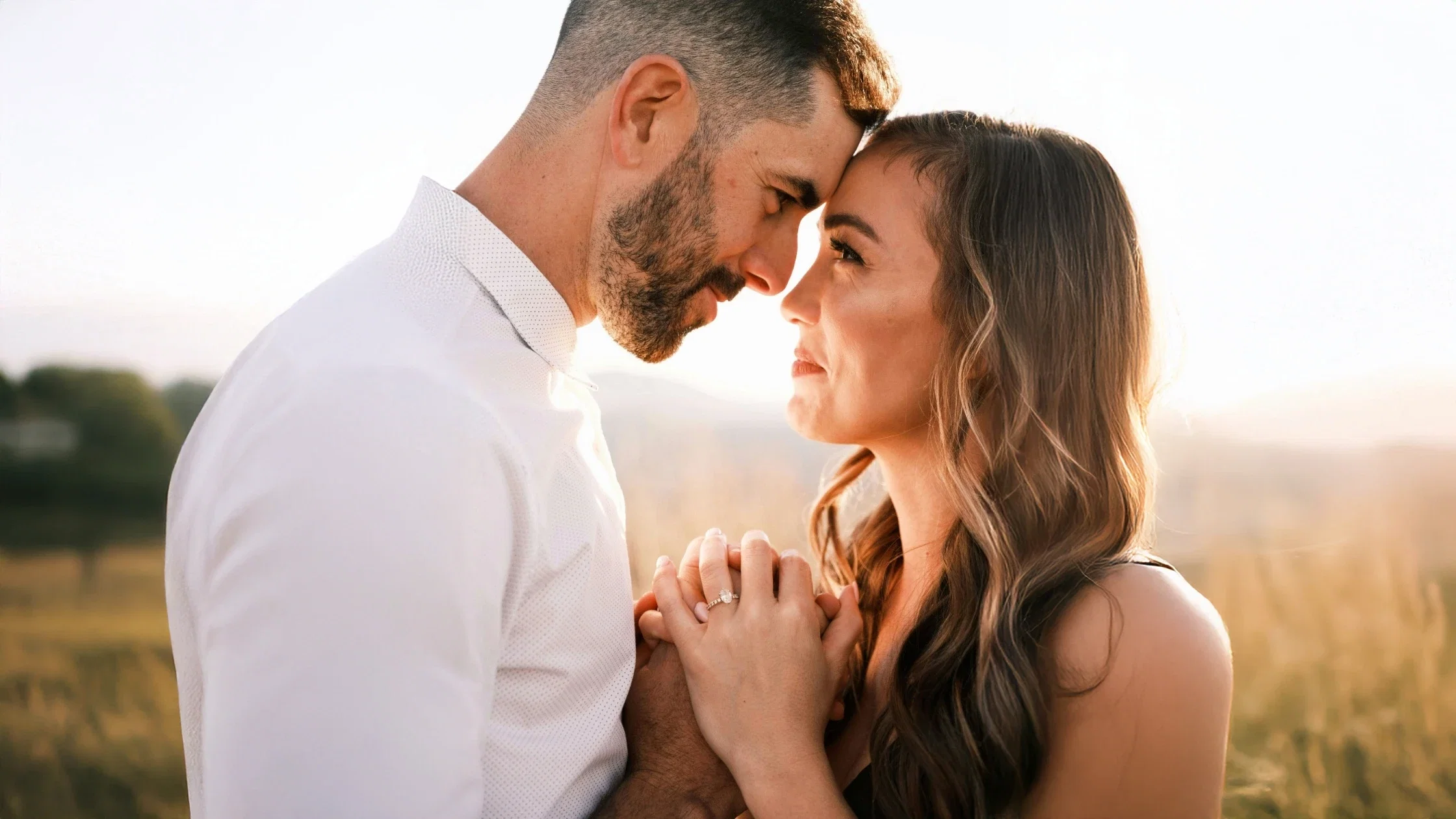 Romantisch koppel staat dicht tegen elkaar aan tijdens zonsondergang in een open veld en houdt elkaars handen vast met zichtbare verlovingsring.
